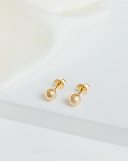 Close-up of polished ball stud earrings with a shiny gold finish, highlighting their 18k gold plating. The earrings have a simple, minimalistic design, perfect for both first and second piercings.