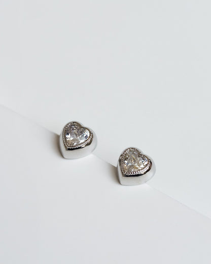  Close-up of heart-shaped earrings featuring clear zirconias set in rhodium-plated metal, showcasing a shiny, elegant finish.