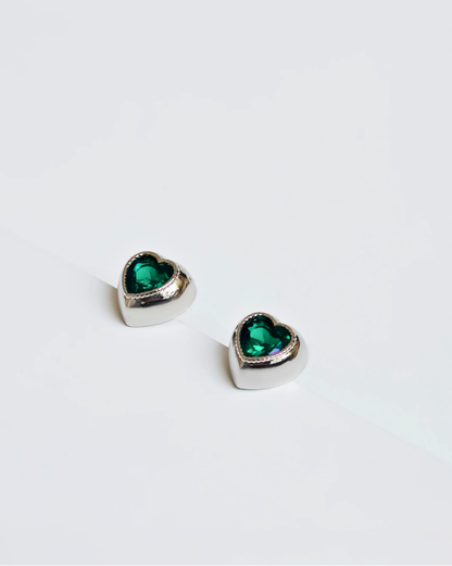  Close-up of heart-shaped earrings featuring vibrant green zirconias set in rhodium-plated metal, showcasing a shiny, elegant finish.