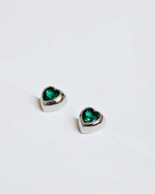  Close-up of heart-shaped earrings featuring vibrant green zirconias set in rhodium-plated metal, showcasing a shiny, elegant finish.